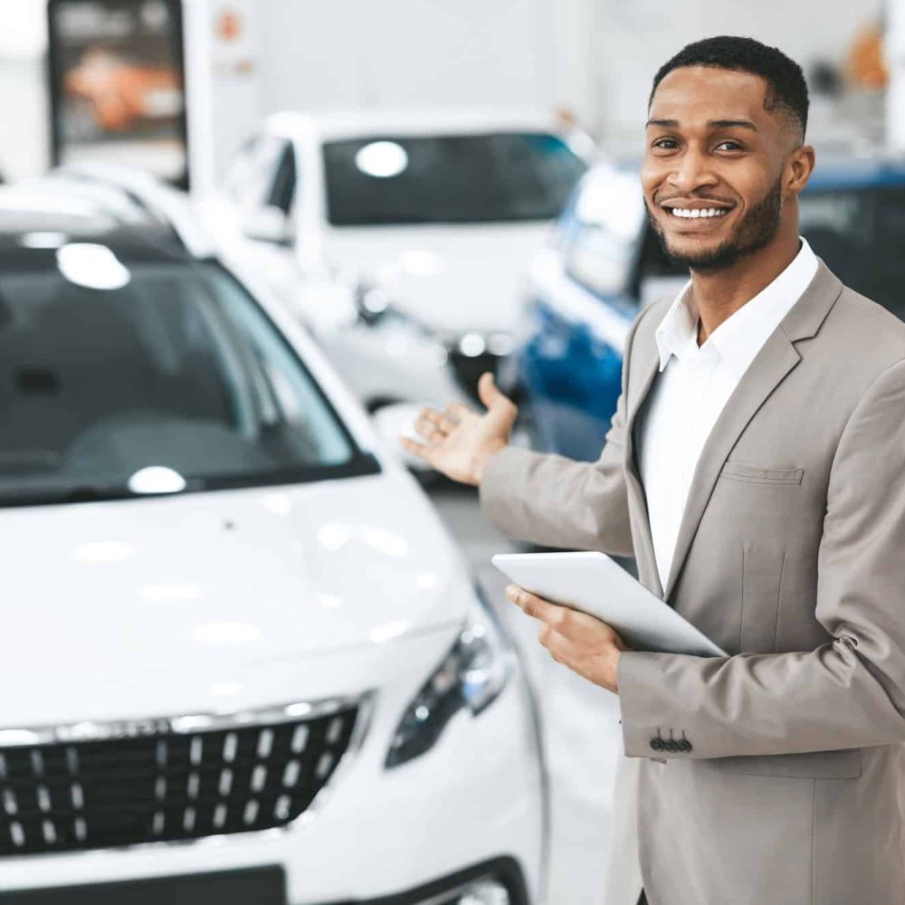 cheerful-car-dealer-gesturing-showing-brand-new-automobile-standing-indoors.jpg